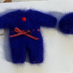 Peut inclure: Un ensemble miniature bleu cobalt duveteux comprenant une combinaison à manches longues et jambes, ainsi qu'un chapeau assorti. La combinaison est ornée d'un bouton rouge et d'un nœud en ruban rouge et blanc à la taille. L'ensemble est probablement pour une poupée ou un petit jouet.