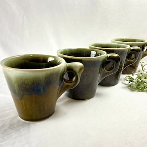 May include: Four ceramic mugs with a dark green and brown glaze. Each mug has a rounded handle and a flared shape. The mugs are arranged in a row, with a small bouquet of white flowers to the right.