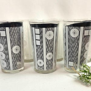 May include: A set of three clear glass tumblers featuring a black and white graphic pattern of vertical textured stripes, geometric squares, and starburst motifs. The glasses are arranged in a row, with a small cluster of white flowers in the foreground.