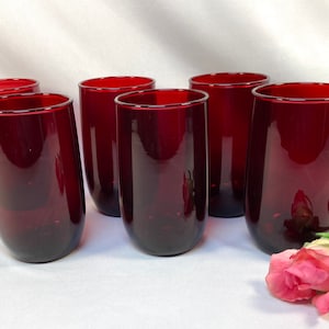 May include: Set of six tall, stemless drinking glasses in a deep red hue. The glasses have a rounded base and a straight, slightly flared rim. A pink rose with green leaves is in the foreground.