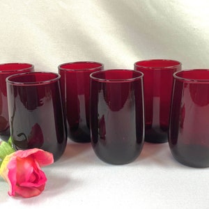 May include: A set of six deep red glass tumblers with a smooth, cylindrical shape and rounded bases. The glasses are arranged in two rows, featuring a consistent dark ruby hue throughout the glass material, accompanied by a single artificial pink rose.