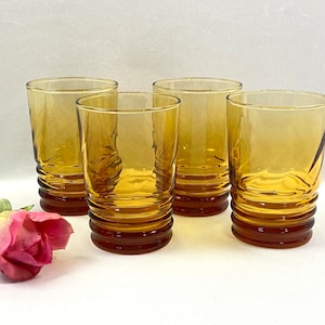 May include: A set of four amber-colored glass tumblers featuring a ribbed base design with horizontal ridges. Each glass has a smooth upper section and a textured lower portion. A single artificial pink rose with green leaves is placed nearby.