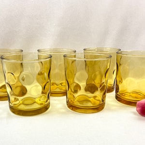 May include: A set of five amber-colored glass tumblers featuring a raised circular dot pattern on the exterior. The glasses are arranged in a group, and a single pink rose with green leaves is positioned to the right of the glassware collection.