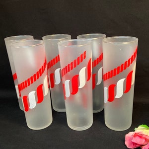 May include: Set of six frosted glass tumblers. Each tumbler features a red and white design. The glasses are cylindrical and appear to be vintage. The background is black.