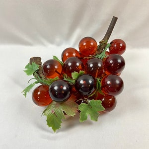 May include: A decorative cluster of grapes with amber-colored glass orbs. The grapes are attached to a brown stem with green leaves. The grapes vary in color from light amber to a deep, dark brown.
