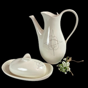 May include: Cream-colored ceramic pitcher and covered dish set. The pitcher has a long spout and a curved handle. Both pieces feature a black geometric design. A small bouquet of white flowers sits nearby.