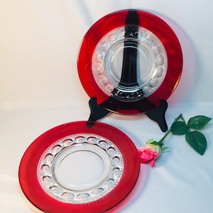 May include: Two red glass plates with clear centers and decorative edges. The plates are displayed on black stands, with a pink rose and green leaves in the foreground. The plates have a circular design.