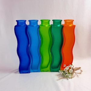 May include: Five colorful glass vases in shades of blue, green, and orange. Each vase has a unique wavy design. A small bundle of white flowers with a twine bow rests in front of the vases.