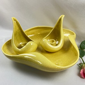 May include: A mid-century modern yellow ceramic candle holder set. The set includes a large, organically shaped dish and two matching candle holders. The pieces have a speckled finish and a glossy sheen. A pink rose is in the background.