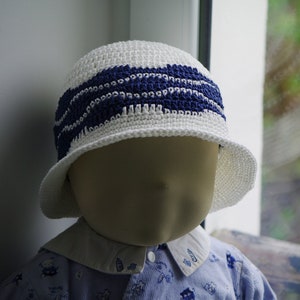 Peut inclure: Chapeau blanc au crochet avec une large bordure et une bande bleu marine. Le chapeau est présenté sur une tête de mannequin. Le chapeau est probablement pour un bébé ou un tout-petit.