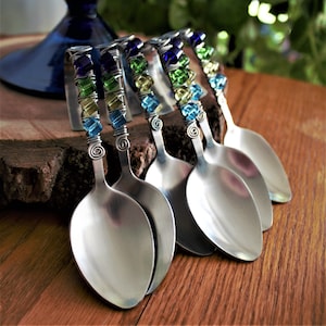 May include: A set of five stainless steel spoons with decorative handles. The handles are wrapped in silver wire and decorated with colourful glass beads in blue, green, and yellow. The spoons are arranged on a wooden surface.