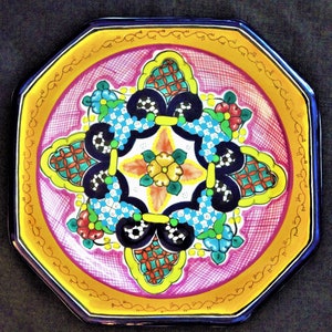 May include: Octagonal ceramic plate with a colorful floral design. The plate features a central flower with four surrounding petals, each with a different pattern. The plate is decorated with yellow, blue, green, red, and white colors.