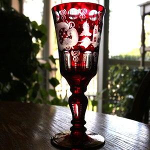 May include: A red glass goblet with a white etched design of a house, trees, and swirls. The goblet has a tall stem and a wide base.