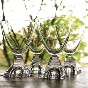 May include: Four clear glass wine glasses with square bases that resemble pyramids. The glasses are arranged in a row on a wooden surface.