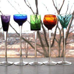 May include: Five colorful, stemmed shot glasses. The glasses are in shades of purple, blue, green, orange, and teal. Each glass has a unique shape and design, with long, slender stems and varying bowl styles. The glasses are arranged in a row.