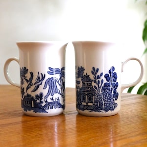 May include: Two white ceramic mugs with blue and white willow pattern designs. The mugs have a handle on the right side.