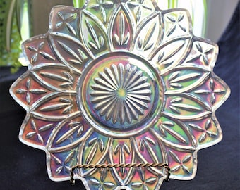 Può includere: Piatto in vetro iridescente con un design a stella. Il piatto ha un bordo smerlato e un motivo centrale in rilievo. I colori variano dall'oro al viola e al verde. Il piatto è esposto su un supporto decorativo in metallo.
