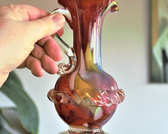 May include: A small, red and clear glass pitcher with a twisted design around the middle. The pitcher has a handle and a round base.