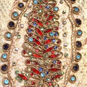 May include: Close-up of a gold fabric with intricate embroidery and embellishments. The design features a central, leaf-like pattern adorned with red, blue, green, and clear gemstones. The edges are outlined with gold thread and additional gemstones.