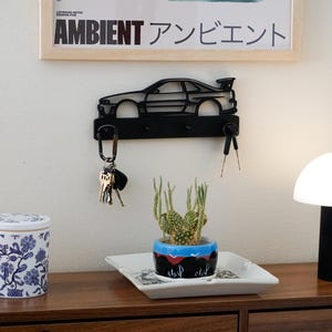 Puede incluir: Un llavero de metal negro con forma de coche, con llaves colgando. Un tarro decorativo con estampado floral azul y blanco, un pequeño cactus y una lámpara con forma de hongo están sobre una superficie de madera.