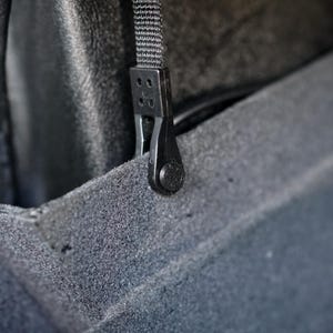 May include: Close-up of a black plastic clip attached to a black woven strap. The clip has a series of holes and a textured button. The strap is connected to a dark gray, textured surface.