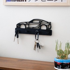 Puede incluir: Llavero de metal negro con forma de coche deportivo. El llavero tiene tres ganchos y está montado en una pared blanca. Las llaves cuelgan de los ganchos. Un pequeño cactus en una maceta azul y negra está sobre una superficie de madera.