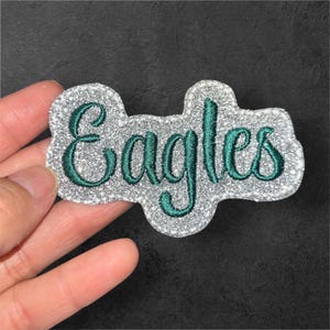 May include: A silver glitter patch with the word "Eagles" embroidered in teal green script. The patch has a unique, cloud-like shape and is likely for apparel or accessories.