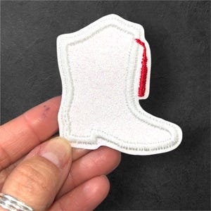 May include: White embroidered patch in the shape of a cowboy boot with a red accent on the side. The boot has a white outline and is held by a hand. The patch is likely for clothing or accessories.