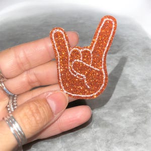 May include: A glittery orange patch in the shape of a hand making the rock and roll gesture. The patch has a white outline and is held by a hand with silver rings. The patch is likely for clothing or accessories.