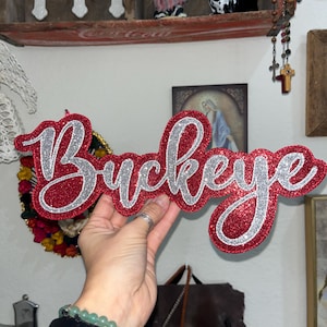 May include: A red glittery "Buckeye" sign with silver glitter trim. The sign is held up in front of a wall with religious art and decorative items.