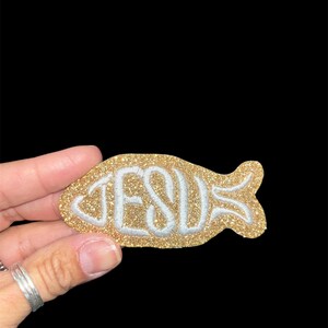 May include: A gold glitter fish-shaped patch with the word "JESUS" in white, outlined in a light blue color. The patch is held against a black background.