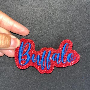 May include: A red glittery patch with the word "Buffalo" embroidered in blue script. The patch is held by a hand against a dark background. The design is likely for apparel or accessories.
