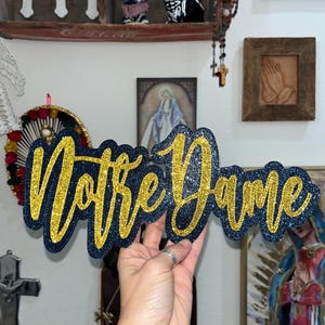 May include: A glittery sign that reads "Notre Dame" in a cursive font. The letters are gold with a dark blue outline. The sign is held up in front of a wall with religious art and decor.
