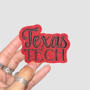Könnte beinhalten: Ein roter, glitzernder Aufnäher mit den Worten "Texas TECH" in schwarzer Schrift. Der Aufnäher wird in einer Hand mit mehreren silbernen Ringen gehalten. Der Hintergrund ist einfarbig hellgrau.
