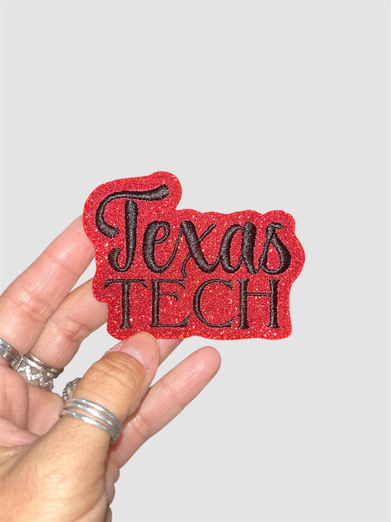 Puede incluir: Un parche rojo brillante con las palabras "Texas TECH" en escritura negra. El parche se sostiene en una mano con m&uacute;ltiples anillos plateados. El fondo es de color gris claro liso.