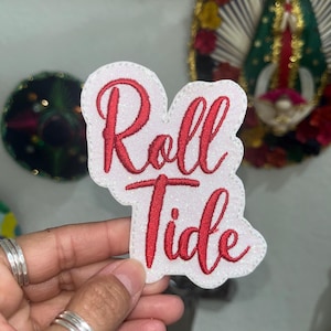 May include: A white glittery fabric patch with red embroidered cursive text that reads Roll Tide. The patch has a die-cut shape that follows the contour of the lettering and features a stitched border around the entire perimeter.