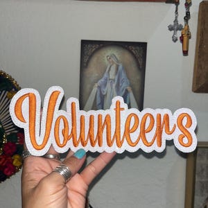 May include: A white embroidered patch featuring the word Volunteers in a cursive orange font with a thick white border. The patch has a textured, glittery finish and is shaped to follow the contours of the lettering.