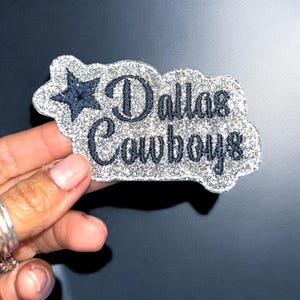 May include: A glittery patch with the words "Dallas Cowboys" in navy blue script. A navy blue star is in the upper left corner. The patch has a silver glitter border and is held by a hand.
