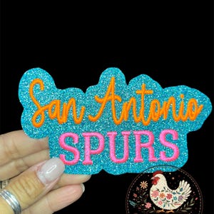 May include: A glittery blue patch with the words "San Antonio SPURS" in orange and pink. The patch is held by a hand against a black background. A logo with a chicken and the words "Cluck & Stitch" is in the lower right corner.