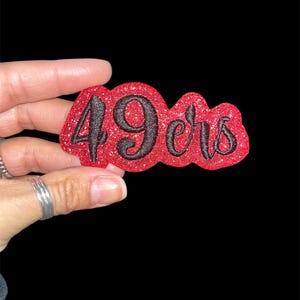May include: A red glittery patch with the text "49ers" in black script. The patch is held against a black background. The patch is likely an emblem for a sports team.