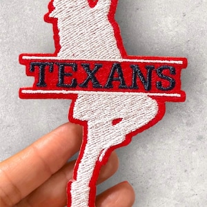 May include: Embroidered patch in the shape of the state of Texas with a red border and the word "TEXANS" in black lettering. The interior of the patch is white with a textured appearance.