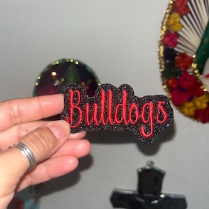 May include: A black glittery fabric patch featuring the word Bulldogs embroidered in red thread. The patch has a scalloped edge and a textured, sparkling surface, with the text written in a cursive, bold font style.