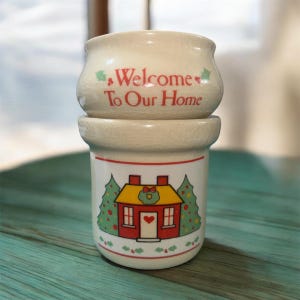 May include: A two-piece ceramic container with a cream-colored, crackled finish. The top piece reads "Welcome To Our Home" in red script. The bottom piece features a red house with a yellow roof, green trees, and a heart. A festive home decor item.