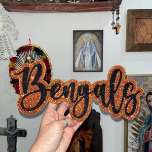 May include: A glittery orange and black sign that reads "Bengals" in a cursive font. The sign is held up in front of a wall with religious art and decorative items. The sign is a decorative item.