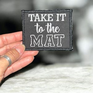 May include: A black square patch with white embroidered text that reads "TAKE IT to the MAT". The patch has a black border and is held by a hand. The patch is likely for clothing or accessories.