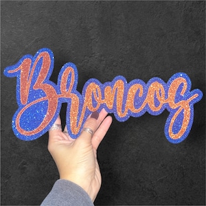 May include: A blue and orange glittery sign that says "Broncos" in a cursive font. The sign is held up against a black background.