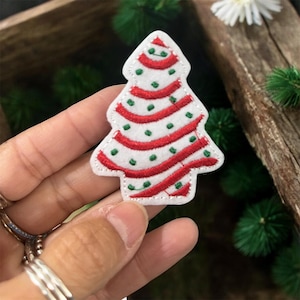 May include: A white Christmas tree-shaped patch with red and green embroidered details. The tree features red horizontal stripes and green dots, with a white border. This festive patch is perfect for holiday-themed crafts and decorations.