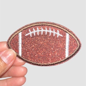 May include: A glittery brown football-shaped patch with white stripes and stitching. The patch has a brown border and is held by a hand against a white background. This patch is suitable for sports-themed crafts.