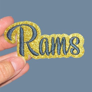 May include: A gold glitter patch with the word "Rams" embroidered in navy blue. The patch has a rounded shape and is held against a solid blue background.