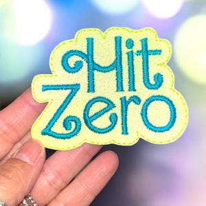 May include: A yellow patch with a wavy border and the words "Hit Zero" embroidered in teal. The patch is held up against a blurred background of blue and white lights.